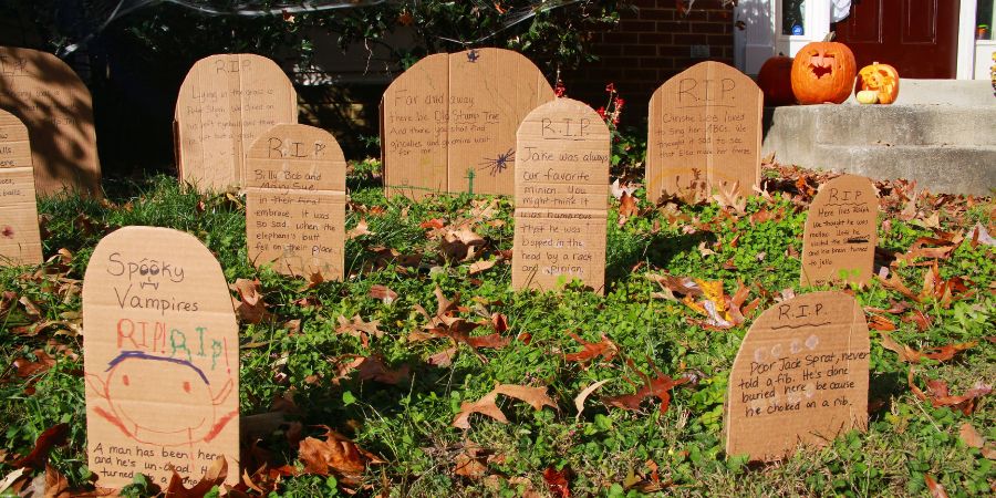 Homemade DIY spooky outdoor Halloween decorations with cardboard tombstones arranged in a front yard graveyard scene beside carved pumpkins.