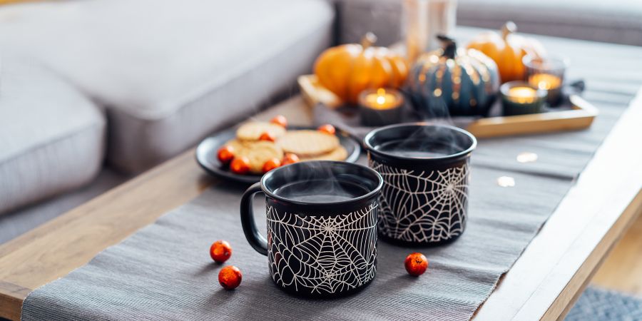 Living room Halloween decorations indoor with black spiderweb mugs, cookies, pumpkins, and candles styled on a cozy coffee table.