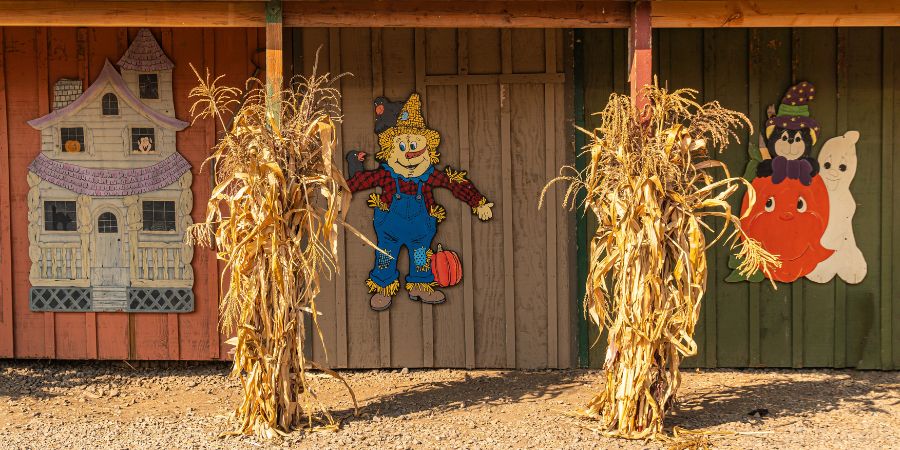 Homemade Halloween decorations outdoors with painted wooden cutouts of a haunted house, scarecrow, pumpkin, and ghost framed by dried corn stalks.