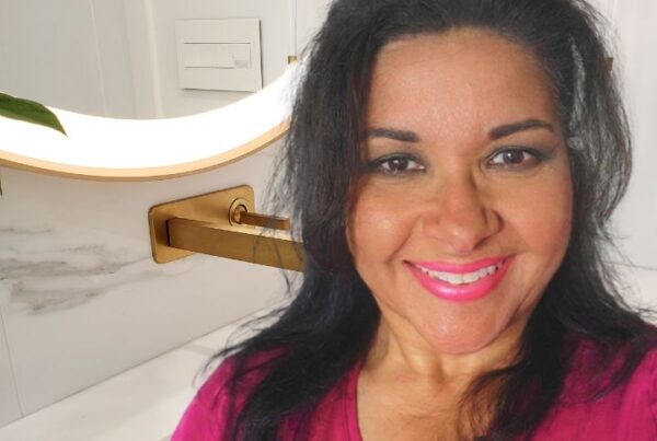 A smiling woman in a pink top pictured in a modern bathroom featuring gold hardware, a white marble vanity, and a round backlit mirror.