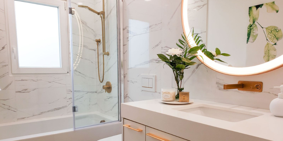 A bright interior design photo of a modern bathroom. In the foreground, a white vanity with slim gold drawer pulls holds an undermount sink and a wall-mounted gold faucet. Above the counter sits a large round LED backlit mirror reflecting a botanical green leaf wall print. A clear vase with a white flower, a candle, and a soap dispenser rest on the vanity top. To the left, a glass-enclosed bathtub and shower combination features white marble-patterned wall tiles and brushed gold shower fixtures.