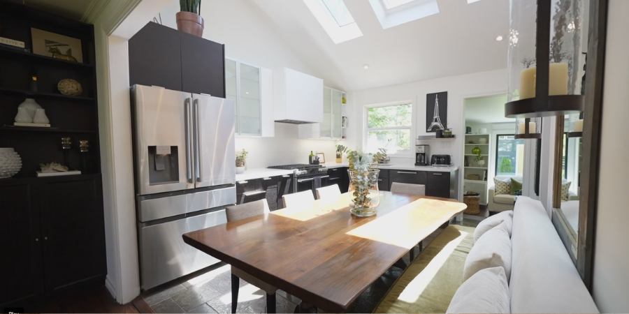 An interior design photo showcasing an open-concept kitchen and dining space illuminated by natural light from vaulted ceiling skylights. In the foreground, a long, natural wood dining table is paired with neutral chairs and a plush white cushioned bench. The background kitchen features dark lower cabinets, white upper cabinets, a white range hood, and a stainless steel French door refrigerator. A black built-in bookcase stands on the far left, and a doorway on the right leads into a brightly lit living area.
