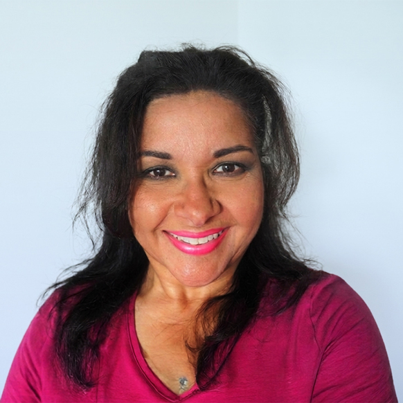 A close-up headshot portrait of a smiling woman with shoulder-length dark hair, wearing bright pink lipstick and a magenta V-neck top, posed in front of a neutral light blue wall.