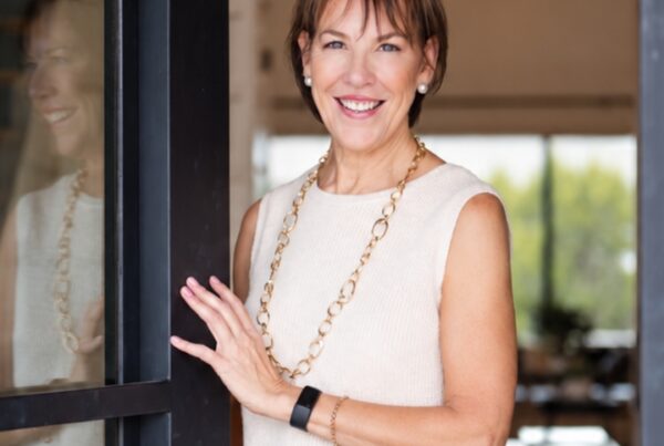 A smiling woman with short brown hair stands in a doorway, gently holding open a dark-framed glass door. She is wearing a sleeveless cream-colored top, a long gold chain necklace, pearl earrings, and a black fitness tracker on her wrist, with a brightly lit interior space visible in the blurred background.