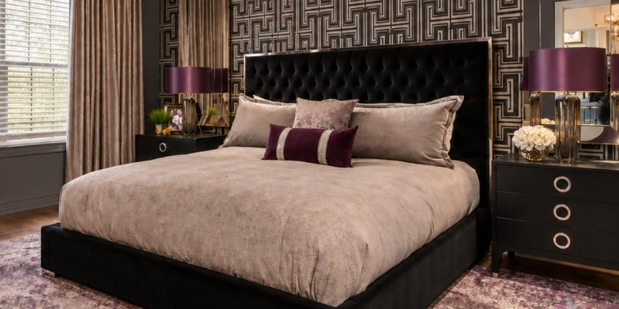 A darkly styled bedroom featuring a black tufted headboard, taupe bedding with dark purple accent pillows, and black nightstands holding lamps with purple shades, all set against a geometric black and metallic wallpaper.