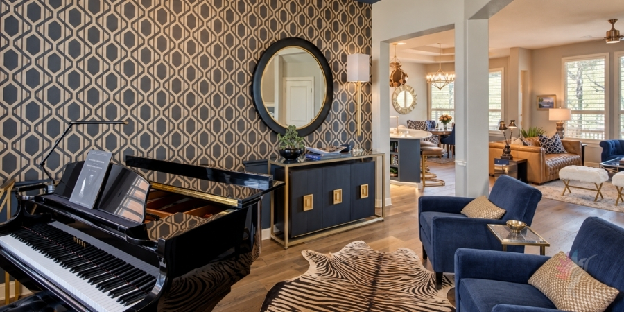 A living area featuring a black grand piano in the foreground, a zebra-print rug, two blue armchairs, and a dark console table under a large round mirror set against geometric patterned wallpaper, with an open view into an adjacent kitchen and living space.