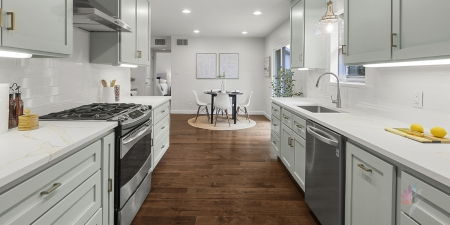 A bright, modern kitchen with light grey shaker cabinets, white countertops, stainless steel appliances, and dark hardwood floors that extend into a dining area with a round black table and white chairs.
