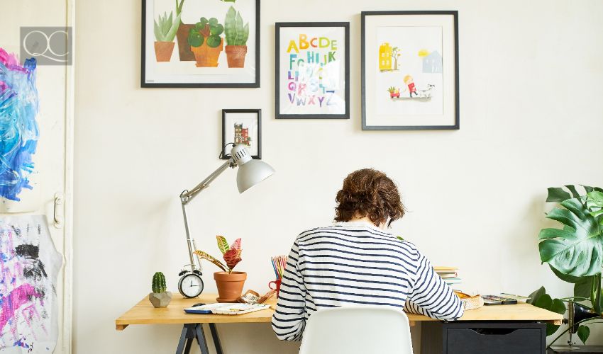 Female artist at her workplace working from home. Young woman dressed in jeans and striped shirt sitting at the table turned backwards. Creating an illustration.