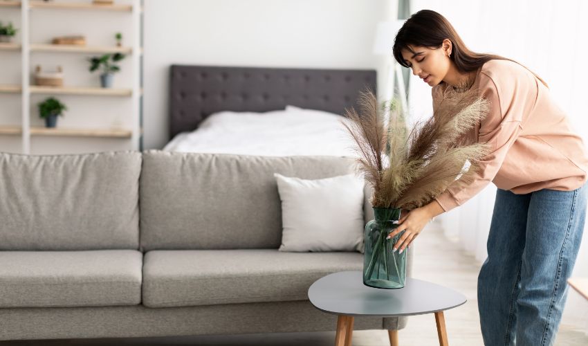 Modern Decoration Concept. Casual young lady putting glass vase with dried flowers on tea table. Millennial woman decorating her modern apartment and bedroom or living room with pampas grass