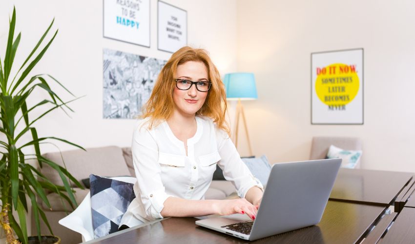 Young woman with long red hair and glasses for view works, prints on a gray laptop keyboard sitting at a wooden table in a bright interior. Subject business, people and technology. Interior design business article.