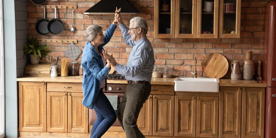 Full length energetic middle aged family couple dancing to disco music in kitchen. Happy old mature man and woman having fun, entertaining together indoors, involved in funny domestic activity. Aging in Place article.