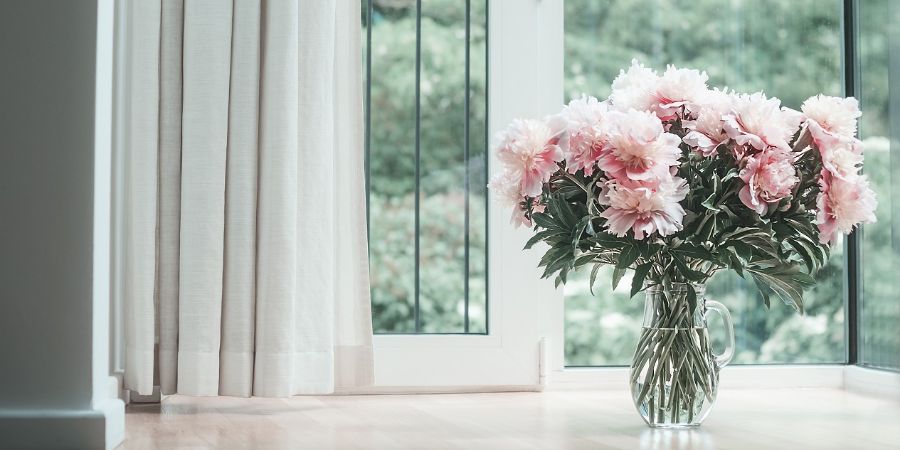 Glorious pastel pink bouquet of peonies in glass jug on floor by window. Flowers in interior design. Cozy home. Virtual design article.