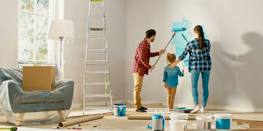 Beautiful Young Family are Showing How to Paint Walls to Their Adorable Small Daughter. They Paint with Rollers that are Covered in Light Blue Paint. Room Renovations at Home. Home renovation living tips.