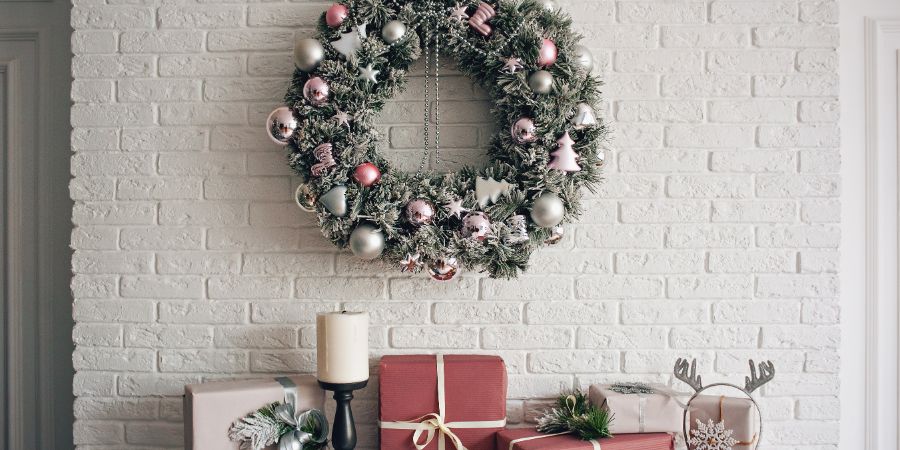 A traditional bright Christmas wreath hanging over the fireplace, on a white brick wall, and packaged gifts are stacked on a fireplace with candles. Christmas concept, new year. Holiday decorating article.