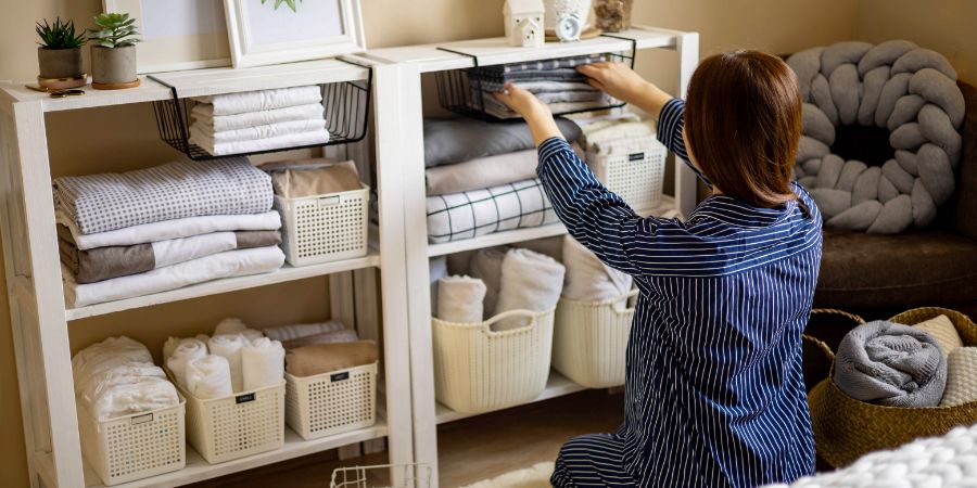 Domestic woman in pajamas neatly putting folded linens into cupboard vertical storage system use Marie Kondo method. Female housewife spring seasonal cleaning space organizer with organized boxes. Organizing mistakes article.